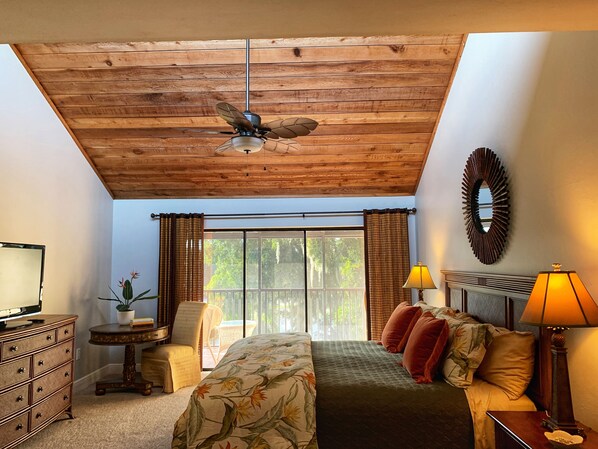 A bedroom with a wooden ceiling, a bed with floral bedding, a nightstand with a lamp, a dresser, a chair, and a window with curtains.