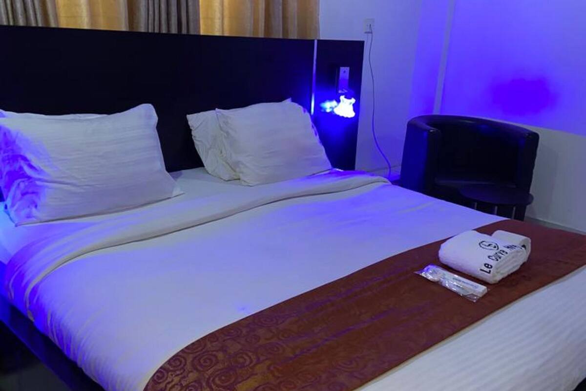 Habitación de hotel con cama, ropa blanca, una colcha roja estampada, una toalla doblada y un televisor sobre un soporte.
