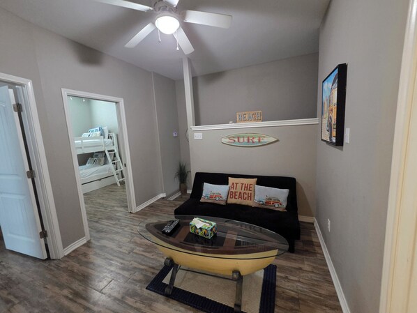 A living room with a glass coffee table, a ceiling fan, a couch with beach-themed pillows, and a wall sign that says 'SURF'.