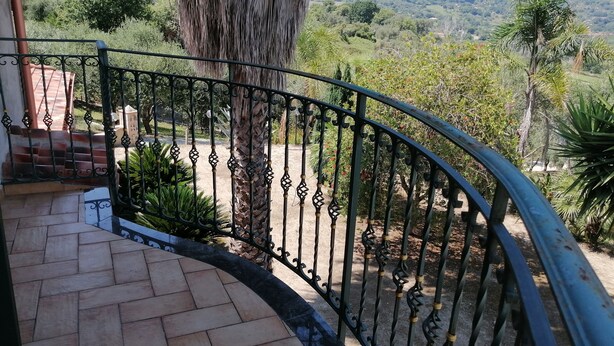 A balcony with a wrought iron railing, a tiled floor, and a view of a lush landscape with trees and hills in the background. - Villa Rosetta in the middle of nature, surrounded by palm and olive trees  (Pizzo)