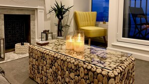 A living room with a fireplace, a yellow armchair, a glass coffee table with candles, and a view of the outside through a window. - Estuary Views Penthouse 🖤 Beachfront with Parking (Southend-on-Sea)