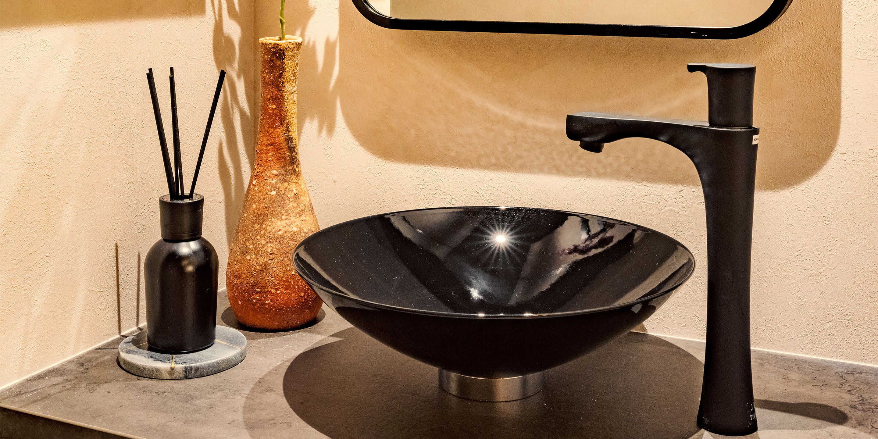Un lavamanos moderno con lavabo negro y grifo, un jarrón alto con una planta y un recipiente con difusores de caña.