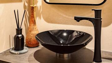 Un lavamanos moderno con lavabo negro y grifo, un jarrón alto con una planta y un recipiente con difusores de caña.