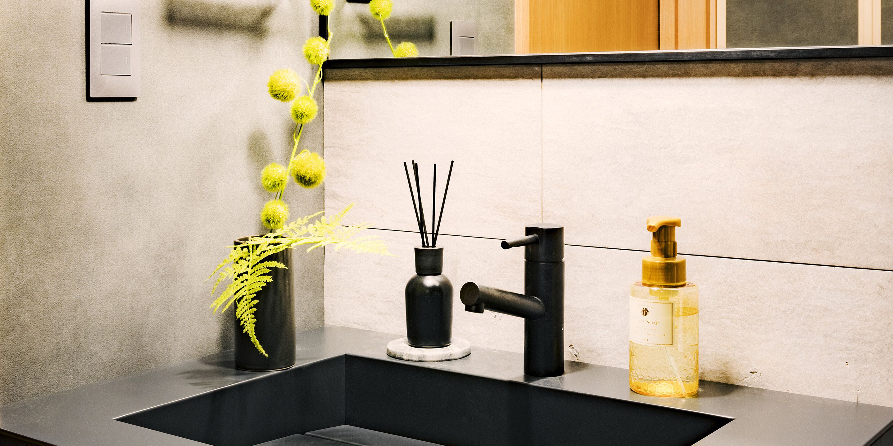 A modern bathroom sink with a black faucet, a small black vase with green foliage, and a bottle of liquid soap.