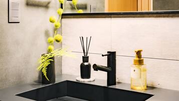A modern bathroom sink with a black faucet, a small black vase with green foliage, and a bottle of liquid soap.