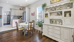 Un coin repas avec une table et des chaises blanches, une cuisine avec des meubles blancs et une étagère décorative agrémentée de plantes.
