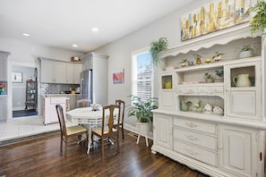 Uma área de refeições com uma mesa e cadeiras brancas, uma cozinha com armários brancos e uma estante decorativa com plantas.