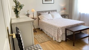 Une chambre à coucher avec un lit, une table de chevet, une lampe, une chaise, une petite table, un tableau au mur et une fenêtre avec des rideaux.