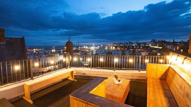 Un espace extérieur aménagé avec des bancs et des tables en bois, offrant une vue sur le paysage urbain et sur une tour imposante.