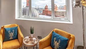 Une pièce avec deux fauteuils jaunes, chacun agrémenté d’un coussin bleu orné d’un motif de homard, une petite table ronde avec une plante et deux tasses, et une vue sur un paysage urbain à travers une grande fenêtre.