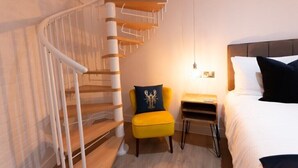 Une chambre d’hôtel moderne avec un escalier en colimaçon, un fauteuil jaune, une table de chevet et un lit avec des oreillers blancs et foncés.