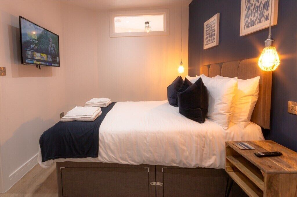 Ein ordentlich bezogenes Bett mit einer weißen und dunkelblauen Tagesdecke, eine Fernbedienung auf einem Holznachtkästchen und ein an der Wand montierter Fernseher.