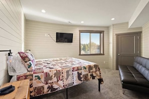 Un dormitorio con cama, televisor montado en la pared, ventana con persianas, puerta y sofá.