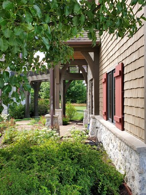 A covered outdoor area with stone pillars and a wooden pergola, surrounded by lush greenery and a stone wall.