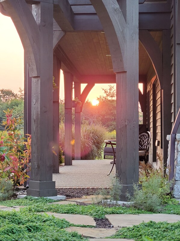 A covered outdoor area with wooden pillars, a pathway leading to a seating area, and a view of the sunset.