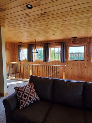 A wooden cabin interior with a sofa, a staircase, and large windows.