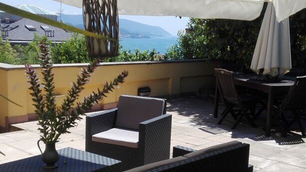 Une terrasse extérieure aménagée avec des meubles en osier, une table ornée d’un vase et une vue sur la mer et les montagnes.