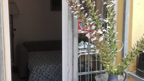 Une porte ouverte donne sur une chambre à coucher où l’on aperçoit une couvre-lit à motifs, une plante en pot posée sur une commode en bois et une fenêtre ornée de barreaux en métal.