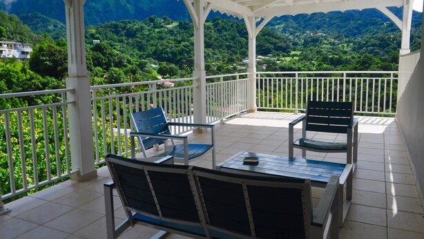 A covered outdoor seating area with chairs and a table, overlooking a lush green landscape and mountains.