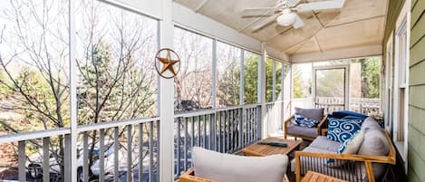A covered porch with wooden furniture, a ceiling fan, and large windows offering a view of trees and parked cars.