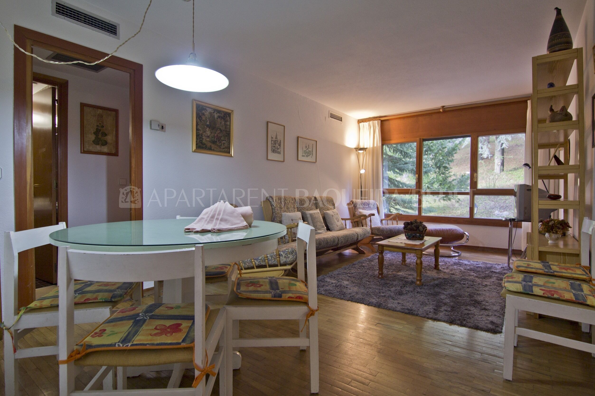 A living room with a glass-top dining table, white chairs, a sofa, and a bookshelf.