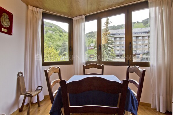 A room with a table and chairs, large windows with curtains, and a view of a building and greenery outside.
