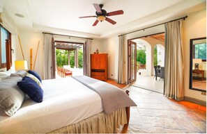 A spacious bedroom with a large bed, a ceiling fan, and a view of a lush garden through the sliding glass doors.