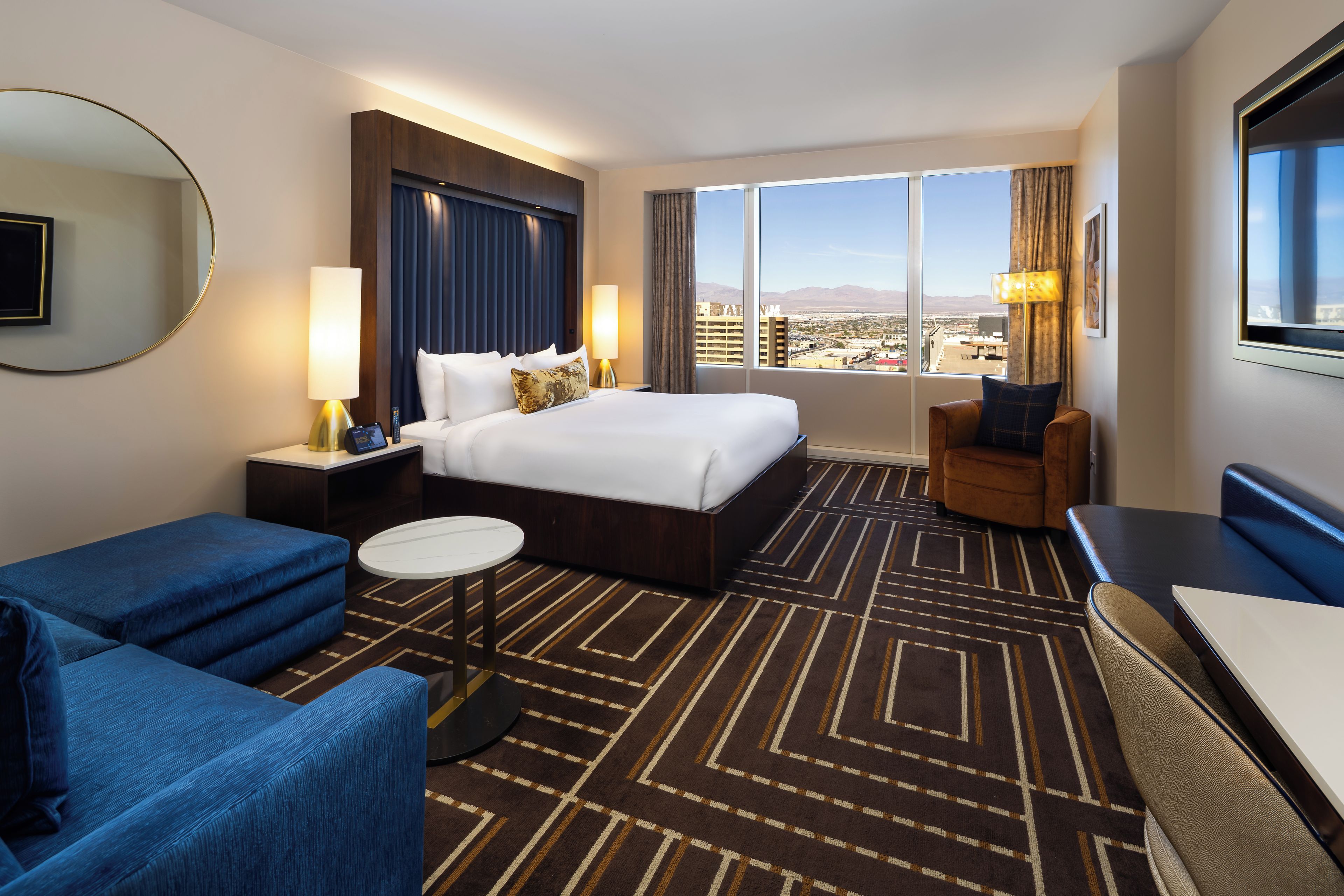 A hotel room with a large bed, a desk, a chair, a round coffee table, and a view of the city.