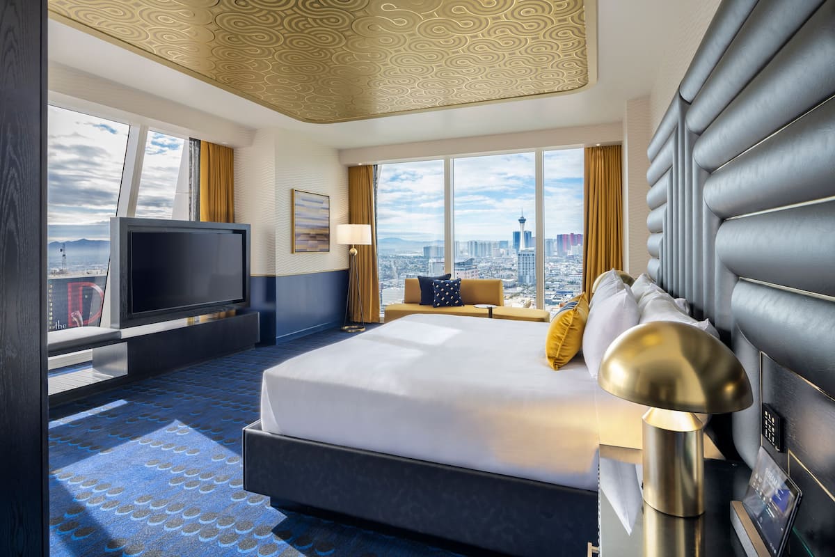 A modern hotel room with a large bed, a flat-screen TV, a city view, and a unique ceiling design.