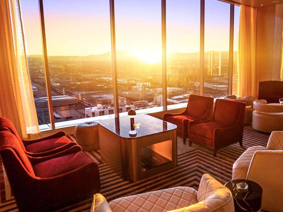 A modern hotel room with a large window offering a city view, a comfortable seating area with red and white chairs, and a small table.