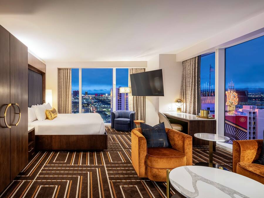A hotel room with a large bed, a desk, a TV, and a view of the city at night.