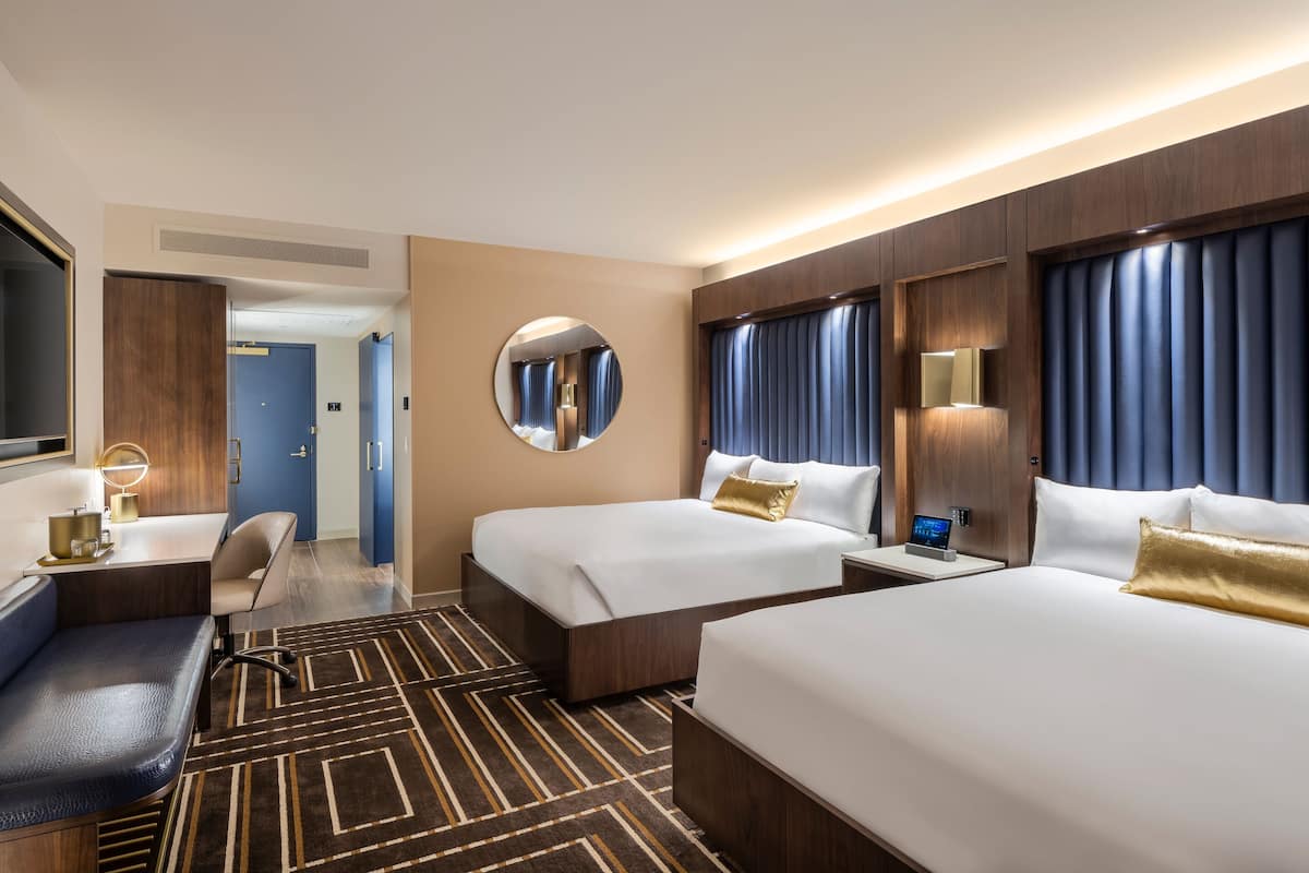 A hotel room with two beds, a desk, a chair, a round mirror, a bedside table with a lamp, and a large window with curtains.