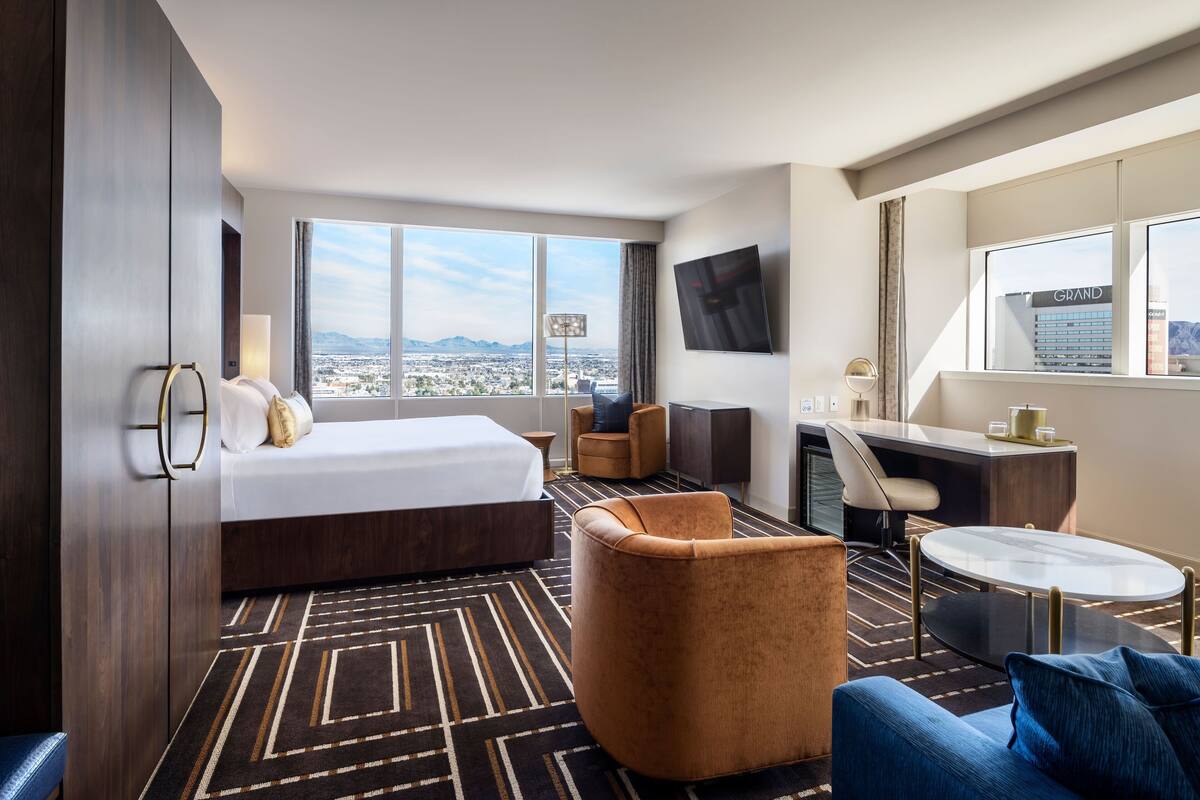A hotel room with a large bed, a desk, a chair, and a sofa. There is a TV mounted on the wall and a window with a view of the city.