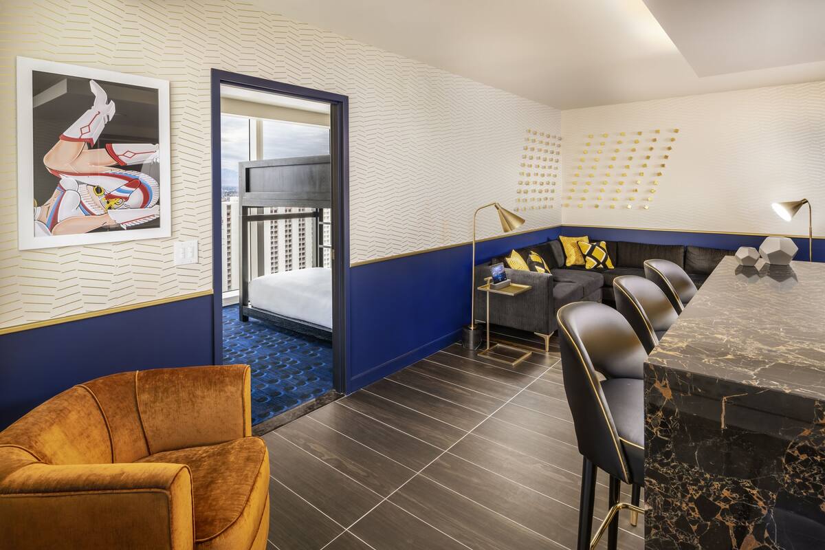 A modern hotel room with a large bed, a seating area with a sofa and chairs, a dining table, and a city view through the window.