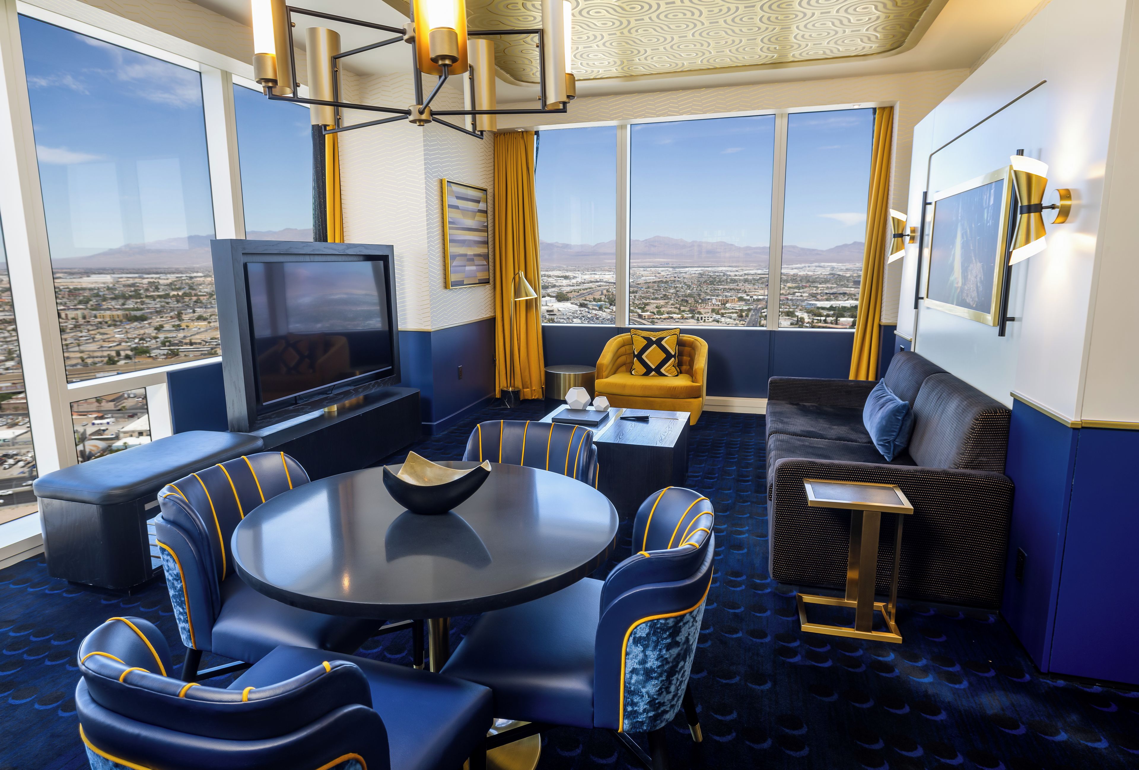 A modern hotel room with a large window, a flat-screen TV, a round dining table, and a sofa.