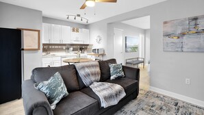 Un salon moderne avec un canapé gris foncé, un ventilateur de plafond et une cuisine en arrière-plan.