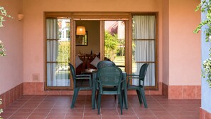 An outdoor patio with a table and chairs, a sliding glass door leading to an interior space, and a view of greenery outside. - Rincón del Golf III - Planta Baja - Beach & Golf (Isla Canela)