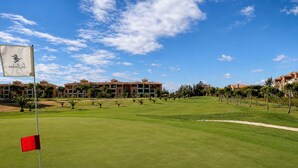 A golf course with a flag, a red flag on the green, and a building complex in the background. - Rincón del Golf III - Planta Baja - Beach & Golf (Isla Canela)