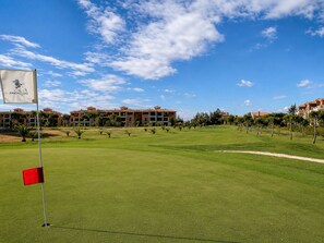 A golf course with a flag, a red flag on the green, and a building complex in the background. - Rincón del Golf III - Planta Baja - Beach & Golf (Isla Canela)