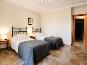 Quarto de hotel com duas camas de solteiro, uma mesa de cabeceira com abajur, um quadro na parede e uma porta de madeira que dá para outro quarto.
