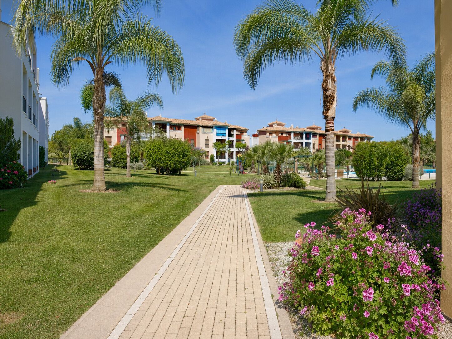 A pathway leading to a building complex with multiple floors, surrounded by well-maintained lawns and palm trees.