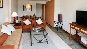 A modern living room with a sofa, coffee table, and television. There is a dining area in the background with a table and chairs. - Rincón del Golf III - Planta Baja - Beach & Golf (Isla Canela)