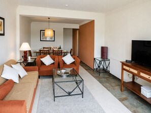 A modern living room with a sofa, coffee table, and television. There is a dining area in the background with a table and chairs. - Rincón del Golf III - Planta Baja - Beach & Golf (Isla Canela)