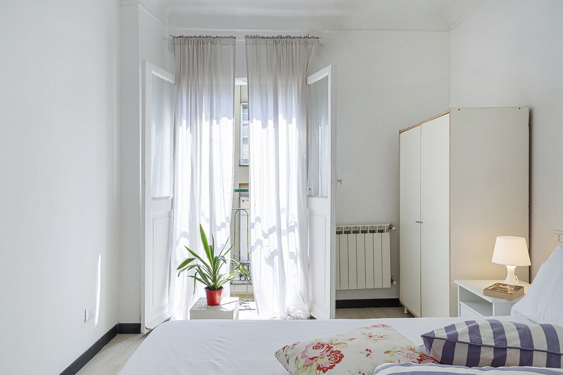Ein Hotelzimmer mit Bett, Nachttisch, Lampe, Spiegel, Heizkörper und einem Fenster mit Vorhängen.