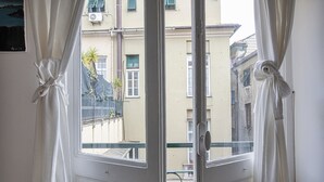 Vista attraverso una finestra con tende bianche ripiegate, che mostra un balcone con ringhiera metallica e un edificio con facciata beige.