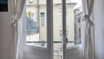 Blick durch ein Fenster mit weißen, zurückgebundenen Vorhängen, der einen Balkon mit einem Metallgeländer und ein Gebäude mit einer beige farbenen Fassade zeigt.