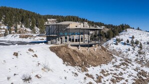 A modern house with a large glass facade, situated on a snow-covered hill with a forested background. - 2 Dome Cabins-1 hr to Yellowstone-Hot tub+Theater+Gaming Room+Pool Table+Air Hockey+Shuffle Board+Secret Kids Playroom (Ashton)