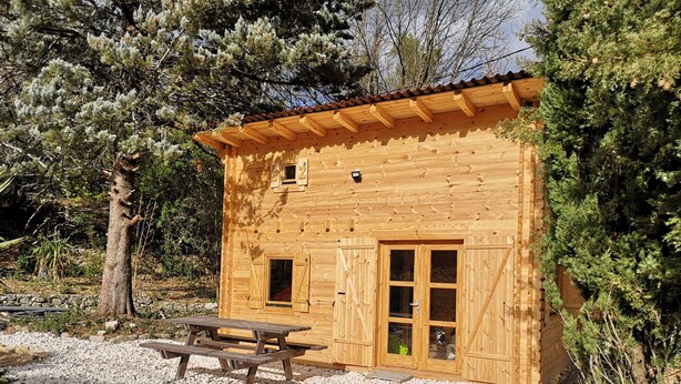Une cabane en bois avec une porte et des fenêtres en bois, une table de pique-nique et un sol recouvert de gravier.