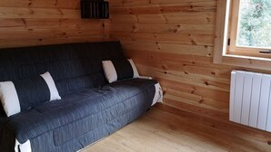 L'intérieur d'un chalet en bois, avec un canapé de couleur sombre adossé à un mur en bois, une fenêtre donnant sur un paysage verdoyant et un radiateur blanc.
