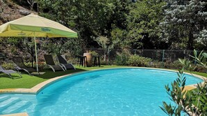 Une piscine avec un parasol vert, entourée d’herbe et d’arbres.